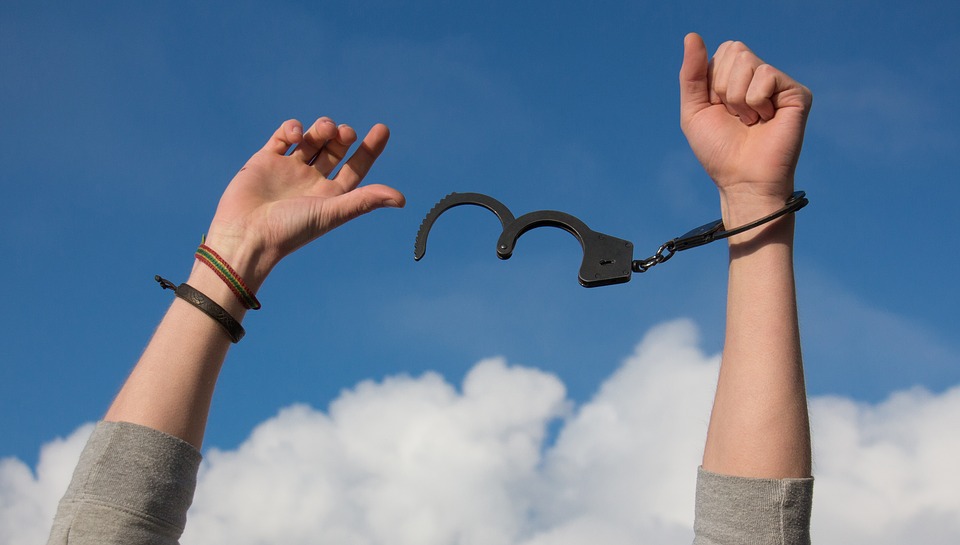 alt="two hands pointing to sky with left hand freed from pair of handcuffs"